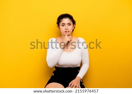 Say no. Indoor portrait of a beautiful woman in stylish clothes covering her mouth. Holds a no-speaking sign and shows shock as he stands against a yellow background. The girl tells the secret