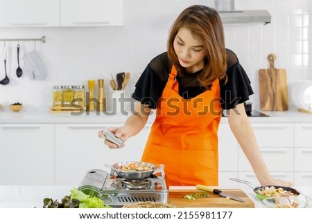 Young Asian mom wearing orange apron learn to cook for family with happy and drop peppers into food on flying pan on gas stove to enhance taste for kid in modern home kitchen