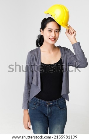 Happy Asian female contractor with yellow protective hardhat standing on gray background in studio and looking at camera