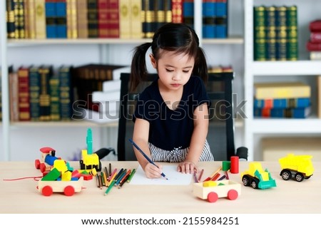 Small pretty pigtails hair preschooler kindergarten happy girl sit on chair drawing cartoon with colored pencils on paper on table full of plastic truck toys in living room at home in front bookshelf.