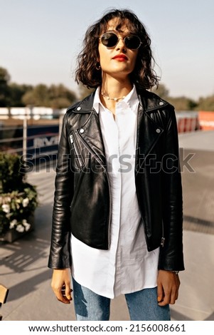 Close-up of cute european young girl looking at camera on blurred background. Brunette with loose short hair wears white shirt, jeans and dark jacket is standing on summer terrace. 