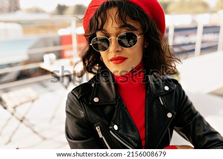 Close up portrait of lovable cute lady with red lips and wearing sunglasses is smiling while waiting for friends in open air cafeteria. High quality photo