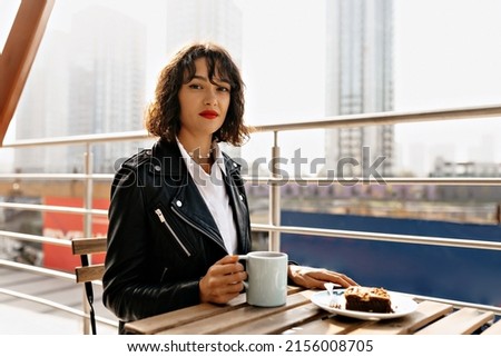 Adorable stylish lady with short wavy hairstyle wearing black jacket and white shirt is sitting on summer terrace with coffee and dessert in sunlight in good mood. High quality photo