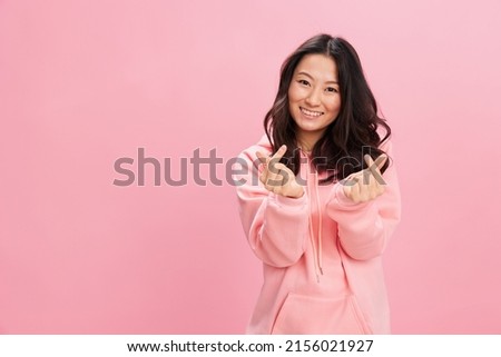 Lovely happy Asian student young lady in pink hoodie sweatshirt show heart shaped sign smiles at camera posing isolated on over pink studio background. Good offer. Fashion New Collection concept