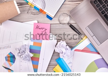 Sticky note with hurry up handwriting and office accessories. White desk background.