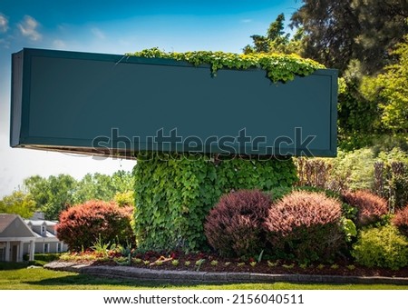 Large advertising sign -blank and copy space - on corner landscaped with vines and flowerbeds with trees and buildings in background