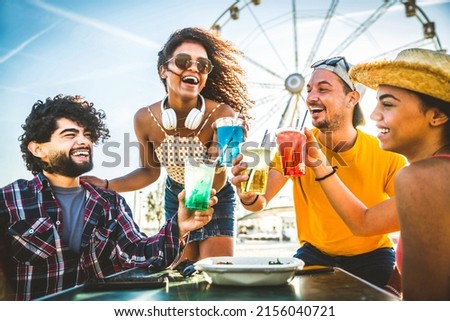 Happy young people cheering cocktail glasses together at beach party - Multi-ethnic friends enjoying happy hour sitting at bar table - Youth lifestyle and summertime vacations concept - Focus on eyes Royalty-Free Stock Photo #2156040721