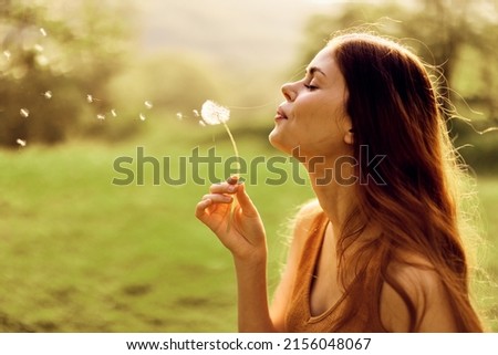 A young woman holds a dandelion in her hands and blows on it, the seeds of the dandelion fly through the air to grow new flowers. Caring for the Earth's Ecology Royalty-Free Stock Photo #2156048067