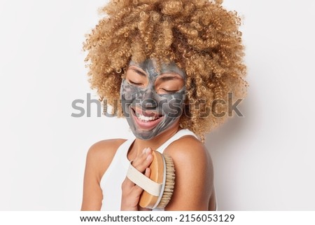 Studio shot of pretty cheerful woman with blonde curly hair applies facial clay mask keeps eyes closed from satisfaction uses body massager poses against white background. Skin care concept.