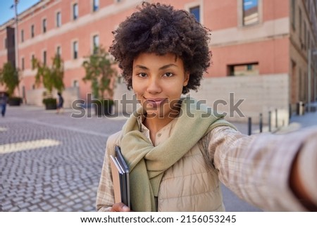 Outdoor shot of beautiful woman student with dark curly hair dressed in casual clothes holds digital tablet and notepads keeps arm outstretched poses at city street during sunny day goes to university