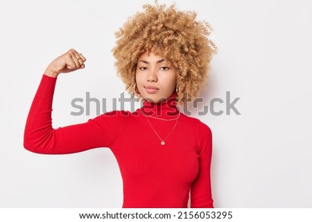 Portrait of self confident curly haired woman flexes biceps shows arm muscles looks seriously at camera wears red turtleneck poses against white background has workout. She can stand up for herself Royalty-Free Stock Photo #2156053295