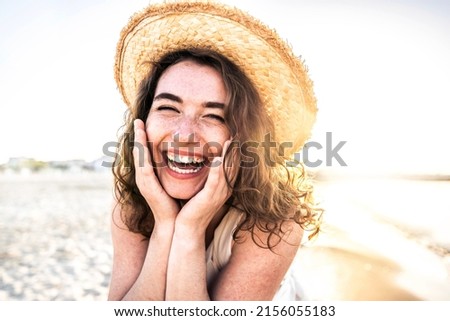 Young joyful woman in white shirt wearing hat smiling at camera on the beach - Traveler girl enjoying freedom outdoors on a sunny day - Wellbeing, healthy lifestyle and happy people concept