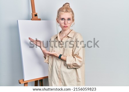 Beautiful caucasian woman with blond hair standing by painter easel stand presenting canva depressed and worry for distress, crying angry and afraid. sad expression. 
