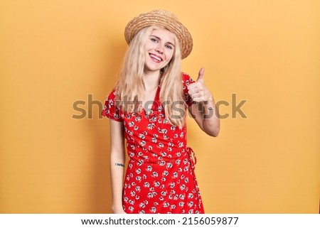 Beautiful caucasian woman with blond hair wearing summer hat doing happy thumbs up gesture with hand. approving expression looking at the camera showing success. 