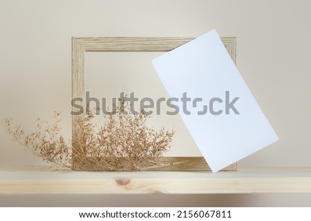 Delicate mockup template with horizontal wooden frame, tilted white card and dry filed plant on wooden background.