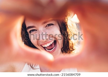 Close up image of smiling woman in swimwear on the beach making a heart shape with hands - Pretty joyful woman laughing at camera outside - Healthy lifestyle, self love and body care concept