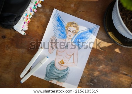 Hand drawn angel on paper and set of alcohol markers on wooden table at art studio.