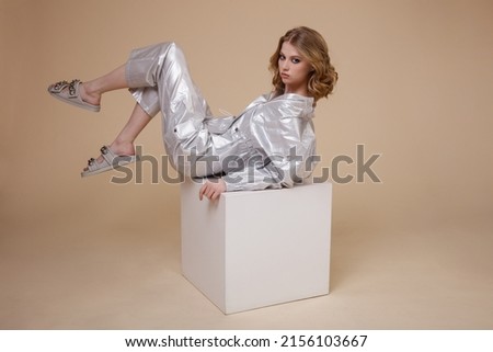High fashion photo of a beautiful elegant young woman in pretty silver platinum metallic suit, jacket, pants on beige brown background. Slim figure. Studio shot. Model is sitting on a white cube