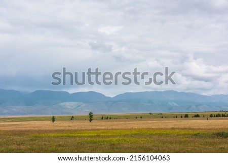 Dramatic alpine view to sunlit steppe and somber large mountains in low clouds during rain. Gloomy mountain landscape with bleak mountain range in rain and steppe in sunlight in changeable weather. Royalty-Free Stock Photo #2156104063