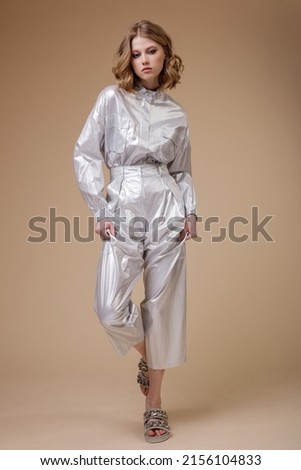 High fashion photo of a beautiful elegant young woman in pretty silver platinum metallic suit, jacket, pants posing on beige coffee background. Slim figure. Studio shot