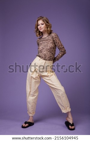 High fashion photo of a beautiful elegant young woman in a pretty beige cream linen trousers, pants, brown blouse with a spotted floral pattern on purple lilac background. Slim figure. Studio Shot