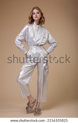 High fashion photo of a beautiful elegant young woman in pretty silver platinum metallic suit, jacket, pants posing on beige coffee background. Slim figure. Studio shot