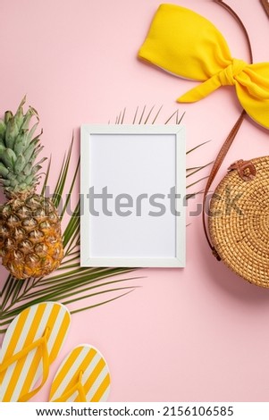 Summer holidays concept. Top view vertical photo of white frame pineapple round rattan bag yellow swimsuit bodice slippers and palm leaves on isolated pastel pink background with copyspace