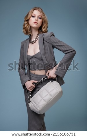 High fashion photo of a beautiful elegant young woman in pretty gray suit, jacket blazer, top, long skirt, handbag, massive chain around the neck posing on blue background. Slim figure. Monochrome