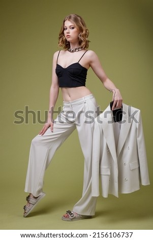 High fashion photo of beautiful elegant young woman in  white, soft gray suit trousers, pants, black top, massive chain around the neck. Jacket blazer on hanger in the hands of model. Green background