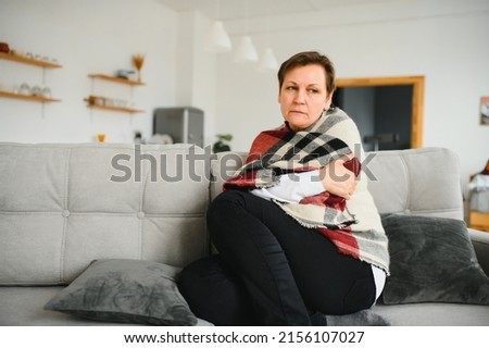 Middle-aged 50s sick frozen woman seated on sofa in living room covered with warm plaid sneezing, runny nose feels unhealthy, seasonal cold, weakened immune system concept Royalty-Free Stock Photo #2156107027
