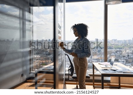 Young African american executive manager businesswoman writing strategy ideas on sticky notes on whiteboard doing business project presentation in contemporary corporate office with panoramic view. Royalty-Free Stock Photo #2156124527