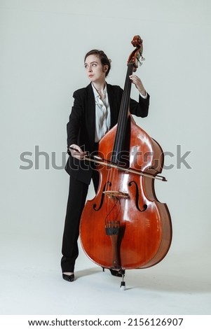Girl musician with long hair in black suit and white shirt plays with the double bass against the white background. Royalty-Free Stock Photo #2156126907