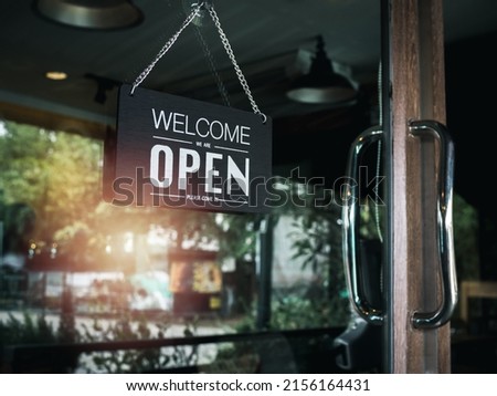 Open sign board with white text on black vintage wooden sign "Welcome we are open please come in" hanging on the glass door of cafe, coffee shop, store or restaurant.