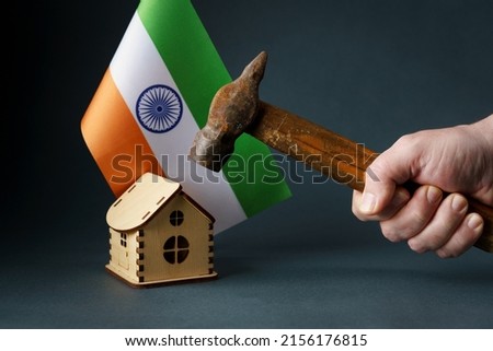 A hammer is raised above the house with the flag of India. An old rusty hammer in a man's hand over a wooden toy house and the flag of India.