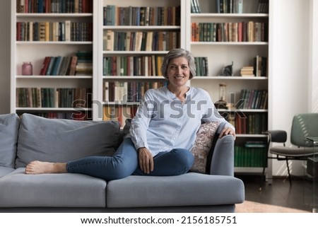Happy positive senior homeowner lady resting on soft comfortable couch at home, looking at camera, smiling, posing in living room interior with bookshelves background