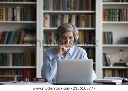Positive focused senior business lady using laptop at table in home office, library with bookshelves, watching learning webinar, online video presentation, thinking over project, work tasks