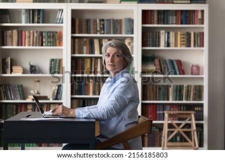 Serious senior university professor woman, mature college student working at laptop in library, looking at camera from workplace with bookshelves in background. Indoor portrait