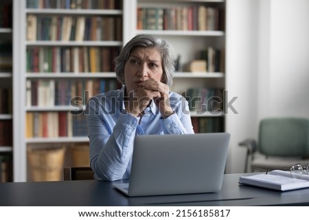 Worried pensive mature business woman using laptop, sitting at table in office with bookshelves, looking away, thinking over challenging tasks, making decision, pondering on problems Royalty-Free Stock Photo #2156185817