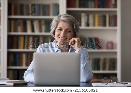 Happy mature businesswoman talking on video call at laptop in office, smiling, laughing, enjoying using online app. Senior student studying in library, watching learning webinar