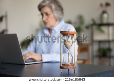 Focused senior gray haired business woman using laptop at hourglass on table, working on project, keeping schedule, completing tasks before deadline. Time management concept Royalty-Free Stock Photo #2156185843