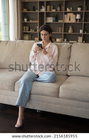 Vertical view serious teenage girl sit on sofa use cellphone chat in social media, share messages, web surfing, search information spend leisure at home. Young gen modern wireless tech overuse concept