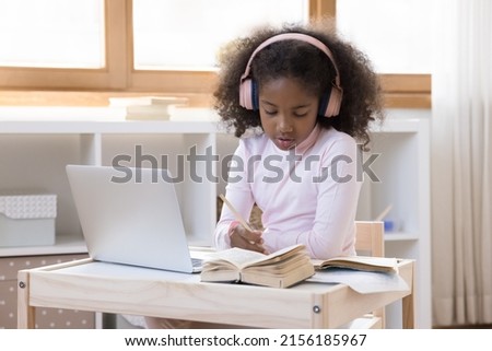 Little African girl in headphones sit at table studying use laptop, e-learning at home. Homeschooling, preschool child busy in preparation for school, do task, gen Z kid learns use modern tech concept Royalty-Free Stock Photo #2156185967