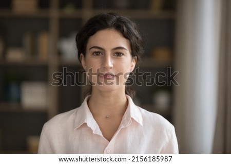 Head shot serious good-looking young 20s woman in casual elegant shirt standing alone in cozy living room staring at camera feels confident pose indoor. Housemaid or real-estate agent portrait concept