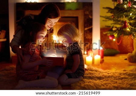 Young mother and her two little daughters opening a magical Christmas gift by a Christmas tree in cozy living room in winter