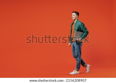 Full size body length side view profile young brunet man 20s wear red t-shirt green jacket hold laptop pc under his hand go move look ahead computer isolated on plain orange background studio portrait
