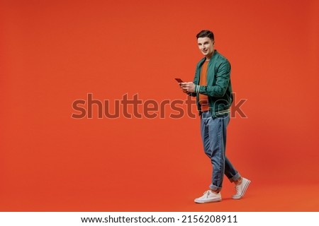 Full size body length side view young brunet man 20s wears red t-shirt green jacket hold in hand use mobile cell phone go step stroll look camera isolated on plain orange background studio portrait.