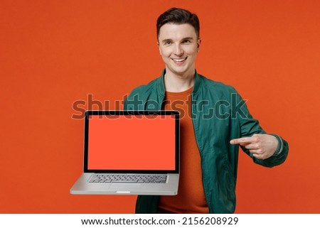 Satisfied young brunet man 20s wear red t-shirt green jacket hold point on laptop pc computer with blank screen workspace area show thumb up gesture isolated on plain orange background studio portrait
