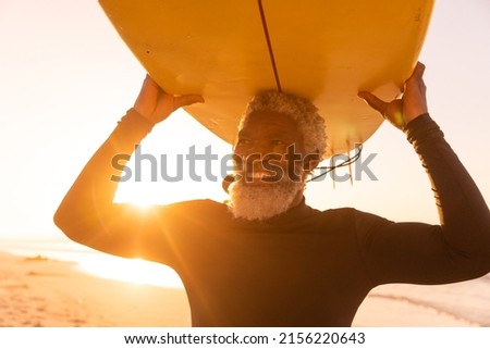 Smiling african american senior man carrying surfboard on head at beach against clear sky at sunset. copy space, sunlight, nature, unaltered, retirement, aquatic sport, holiday and active lifestyle. Royalty-Free Stock Photo #2156220643