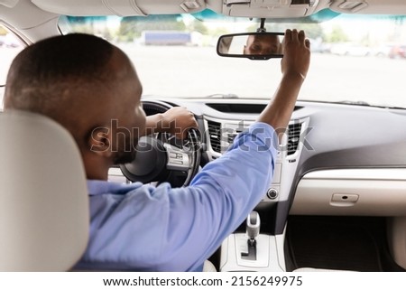 Happy African American Guy Adjusting Rear View Mirror, Enjoying New Auto, Sitting On Driver's Seat In Luxury Vehicle. Man Testing Visibility, Over Shoulder View From The Back Passenger Seat Royalty-Free Stock Photo #2156249975