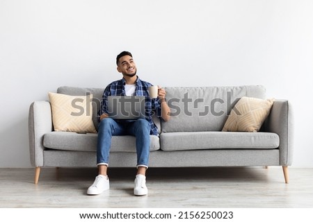 Relax Concept. Happy man drinking coffee using pc sitting on comfortable couch at home in living room and dreaming. Cheerful casual male resting on sofa, enjoying weekend free time, full body length Royalty-Free Stock Photo #2156250023